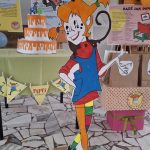 Kolorowa tekturowa wycinanka Pippi Pończoszanki stoi przed tortem i dekoracjami imprezowymi w pomieszczeniu.