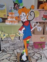 Kolorowa tekturowa wycinanka Pippi Pończoszanki stoi przed tortem i dekoracjami imprezowymi w pomieszczeniu.