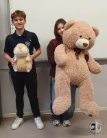 Dwóch uczniów stoi w pomieszczeniu, każdy trzyma dużego pluszowego misia i uśmiecha się do kamery.