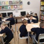 Dzieci siedzą przy stolikach w klasie lub bibliotece, niektóre rozmawiają, a w tle znajdują się półki z książkami i komputery.