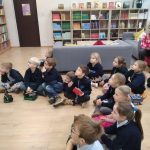 Grupa małych dzieci siedzi na podłodze w bibliotece, uważnie słuchając kogoś poza kamerą.