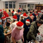 Ludzie w czapkach Mikołaja zebrali się w środku, wykonując rękodzieło przy stołach w świątecznym, ruchliwym otoczeniu.