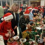 Dzieci i dorośli w czapkach Mikołaja dekorują świąteczne rękodzieła podczas świątecznego wydarzenia w holu.