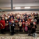 Duża grupa ludzi, wielu w czapkach Mikołaja, pozuje trzymając świąteczne dekoracje i prezenty.