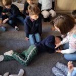 Grupa dzieci przygląda się, jak kobieta demonstruje udzielanie pierwszej pomocy z chłopcem leżącym na podłodze.