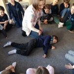 Nauczyciel demonstruje udzielanie pierwszej pomocy grupie dzieci, jedno dziecko leży na podłodze w ramach lekcji.