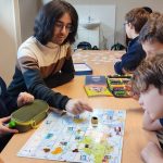 Grupa dzieci i dorosły grają razem w grę planszową przy stole w klasie.