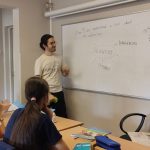 Nauczyciel wyjaśnia lekcję czterem uczniom siedzącym przy biurkach w klasie z tablicą.