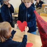 Dzieci w klasie, niektóre w czapkach czarodziejów, wymieniają się dużymi czerwonymi papierowymi sercami przy biurku.