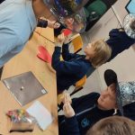 Dzieci w klasie noszą kolorowe kapelusze czarownic i siedzą wokół stołu z przyborami rzemieślniczymi i papierami.