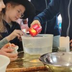 Dzieci w kapeluszach czarownic wlewają czerwony płyn do miski z wodą, przeprowadzając eksperyment naukowy przy stole.