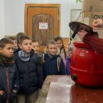 Grupa dzieci obserwuje, jak ktoś otwiera czerwoną doniczkę w pomieszczeniu.