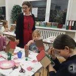 Dzieci malują kartonowe domki przy stole, podczas gdy kobieta w czerwonej sukience nadzoruje zajęcia w klasie.