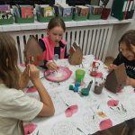 Troje dzieci siedzi przy stole i maluje kartonowe domki, a w tle widać przybory plastyczne i książki.