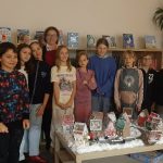 Grupa dzieci i dwoje dorosłych stoi wokół stołu z wystawą zimowej wioski w bibliotece.