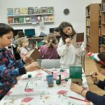 Dzieci malują kartonowe domki przy stole w bibliotece lub klasie, otoczone książkami i przyborami plastycznymi.