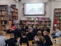 Nauczycielka czyta grupie dzieci w bibliotece; na ekranie za nią wyświetlany jest pokaz slajdów.