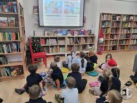 Dzieci siedzą na podłodze w bibliotece, oglądając prezentację na ekranie w pobliżu regałów z książkami i małego drzewka.