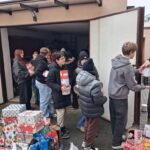 Grupa ludzi podaje puszki z jedzeniem i zapakowane prezenty przed otwartym garażem wypełnionym kartonami.