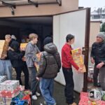 Grupa ludzi przenosi pudełka z prezentami przez drzwi, a obok leżą zapakowane prezenty.