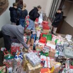 Kilka osób stoi przy stołach pokrytych licznymi zapakowanymi prezentami i torbami na zewnątrz budynku.