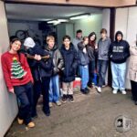 Grupa nastolatków stoi w wejściu do garażu, niektórzy się uśmiechają, jeden ma na sobie czerwony świąteczny sweter z choinką.