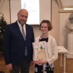 Mężczyzna i dziewczyna stoją w pomieszczeniu; dziewczyna trzyma kolorową książkę i oboje uśmiechają się do kamery.