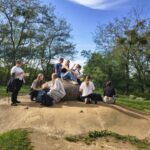 Grupa ludzi siedzi i stoi na dużym kamieniu w parku, otoczonym zielonymi drzewami i trawą.