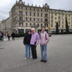 Trzy dziewczyny w zimowych kurtkach stoją razem na placu miejskim z zabytkowymi budynkami w tle.