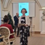 Uśmiechnięta kobieta w czarno-białej sukience spaceruje z książką podczas imprezy w pomieszczeniu, a za nią siedzą ludzie.