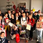 Grupa podekscytowanych dzieci w świątecznych swetrach trzyma prezenty i uśmiecha się w klasie udekorowanej na Boże Narodzenie.