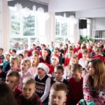 Dzieci w odświętnych zimowych ubraniach i czapkach Mikołaja siedzą razem, oglądając świąteczne wydarzenie w jasnym pokoju.