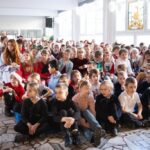 Duża grupa małych dzieci siedzi uważnie w jasnym pokoju, niektóre w odświętnych strojach.