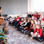 Osoba gra na gitarze dla grupy dzieci w świątecznych strojach, siedzących przy udekorowanej choince.