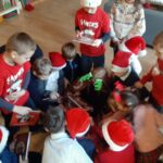 Grupa dzieci w odświętnych strojach i czapkach Mikołaja zebrała się wokół, przeglądając książki na podłodze.