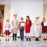 Siedmioro dzieci w odświętnych strojach stoi w rzędzie, czytając gazety na udekorowanej scenie podczas świątecznej imprezy.