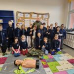 Grupa dzieci i dorosły pozują w klasie z manekinem do resuscytacji i sprzętem medycznym na podłodze.