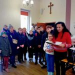 Grupa dzieci i dwoje dorosłych stoi w pomieszczeniu w pobliżu krzyża, niektórzy trzymają dokumenty i prezenty.