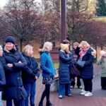 Grupa ludzi, głównie kobiet i dzieci, stoi na zewnątrz w zimowych ubraniach na utwardzonym terenie.