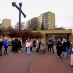 Grupa ludzi gromadzi się w pobliżu dużego krzyża w parku miejskim z budynkami mieszkalnymi w tle.