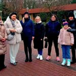 Siedem osób w zimowych ubraniach stoi razem na utwardzonej ścieżce na zewnątrz, uśmiechając się do kamery.