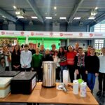 Grupa ludzi stoi za stołami z pojemnikami na żywność i naczyniami w dużej hali.