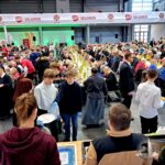 Setki ludzi siedzących przy długich stołach w pomieszczeniach, z wolontariuszami serwującymi jedzenie na dużym wydarzeniu społecznościowym.