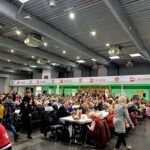 Duży tłum siedzący przy stołach w przestronnej hali, z transparentami i ludźmi serwującymi jedzenie.