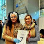 Dwie kobiety w środku, jedna trzymająca tabliczkę z numerem 7, obie uśmiechnięte, ubrane w szaliki i kurtki.