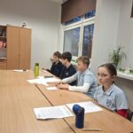 Czterech uczniów w mundurkach siedzi przy stole w klasie z papierami, ołówkami i butelkami wody przed sobą.