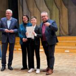 Cztery osoby stoją na scenie; chłopiec w środku trzyma certyfikat i uśmiecha się do kamery.