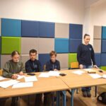 Czterech uczniów siedzi, a jeden stoi przy stole z papierami w klasie z kolorowymi panelami ściennymi.