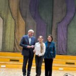 Trzy osoby stoją na drewnianej scenie trzymając papiery, a za nimi znajduje się kolorowa abstrakcyjna kurtyna.