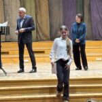 Dziewczyna w białej koszuli schodzi ze sceny, podczas gdy dwoje dorosłych stoi za nią, jeden trzyma papiery, na drewnianej scenie.